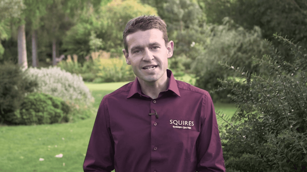 A photo of a man in work uniform standing in a garden and smiling at the camera