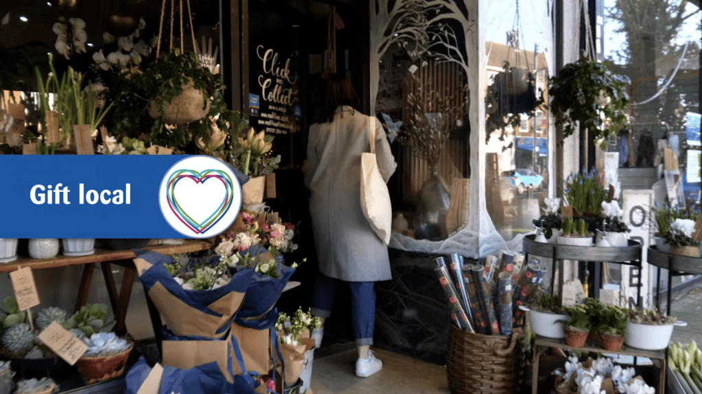 A woman is walking away from the camera through the door of a florist with a large outside display. A small on-screen graphic reads "gift local"