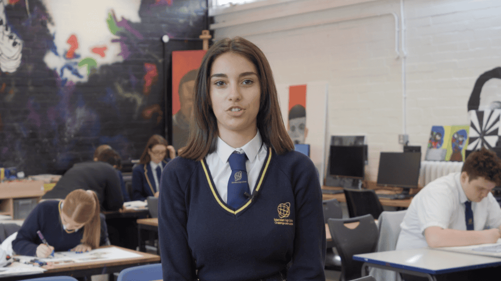Female teenage school student sat on art classroom desk looking to camera with other students working in the background