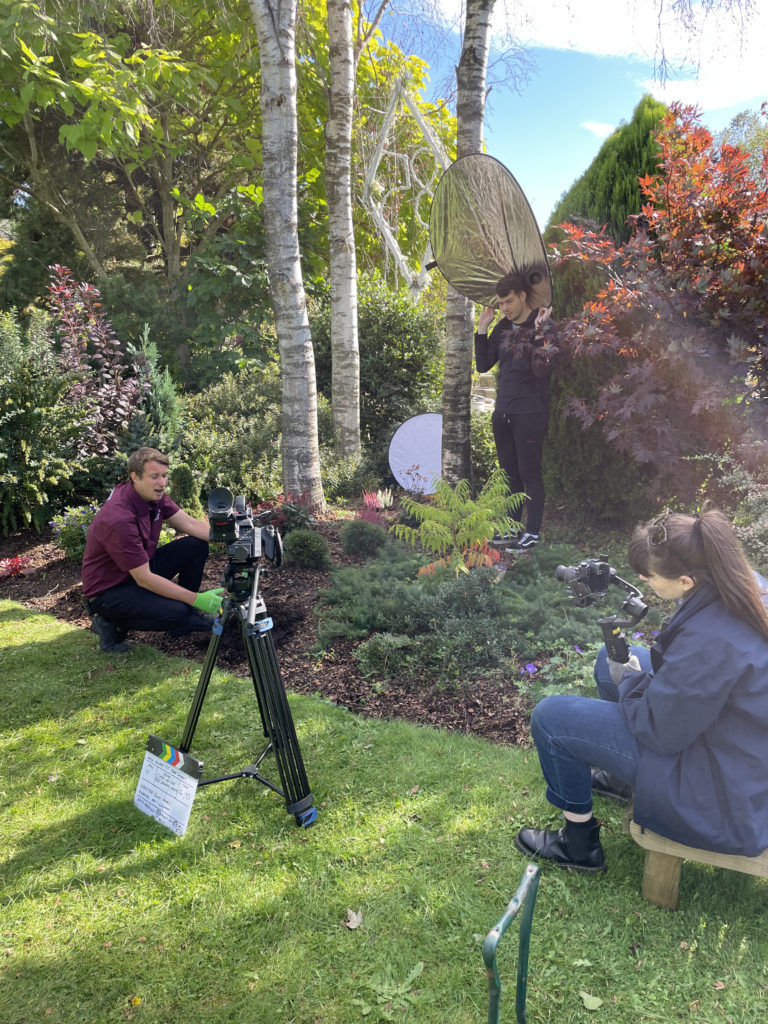 Shot of camera operator and runner holding up a reflector to block sunlight from the presenter