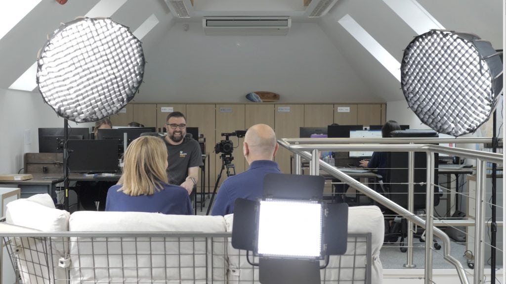 A image from behind the subjects at a shoot showing all of the lights, camera and interviewer