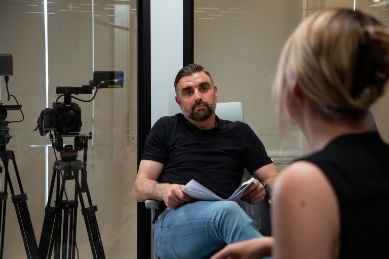 Jovan sitting opposite an interviewee with production schedule on his lap and two cameras over his shoulder